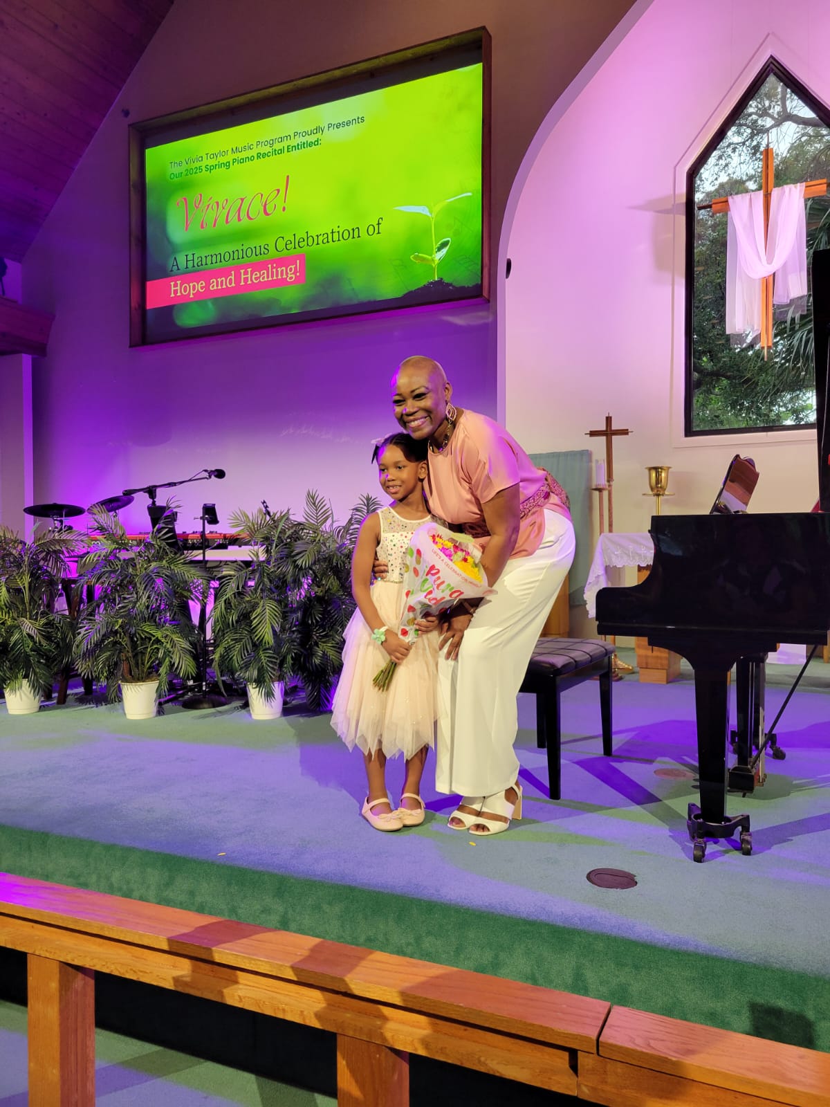 A music student holding flowers stands next to a piano with Vivia Taylor at a recital.