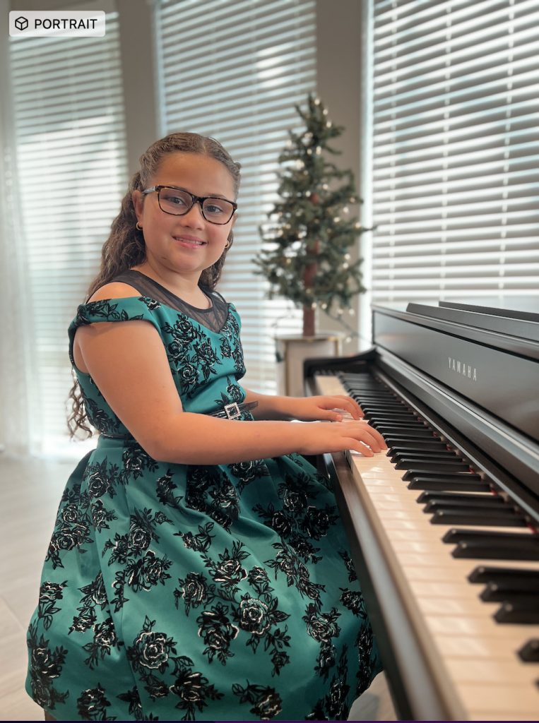 A young music student in a teal dress sits at a piano during a Christmas music recital.