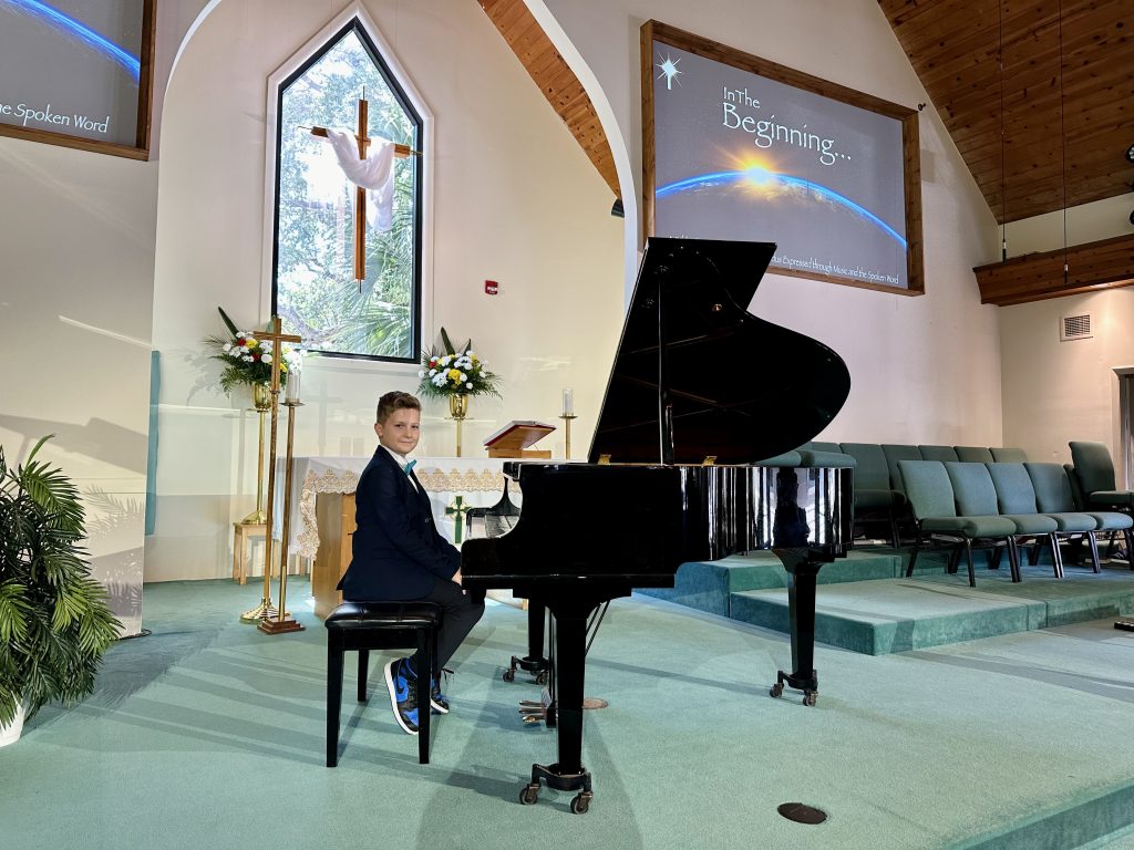 A young music student wearing a suit sits at a piano during a recital in a church