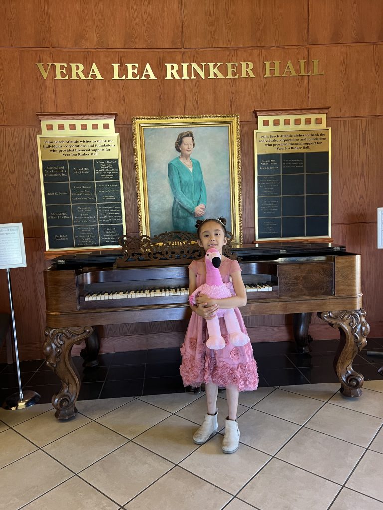 A young music student holding a stuffed flamingo stands in front of an antique piano.