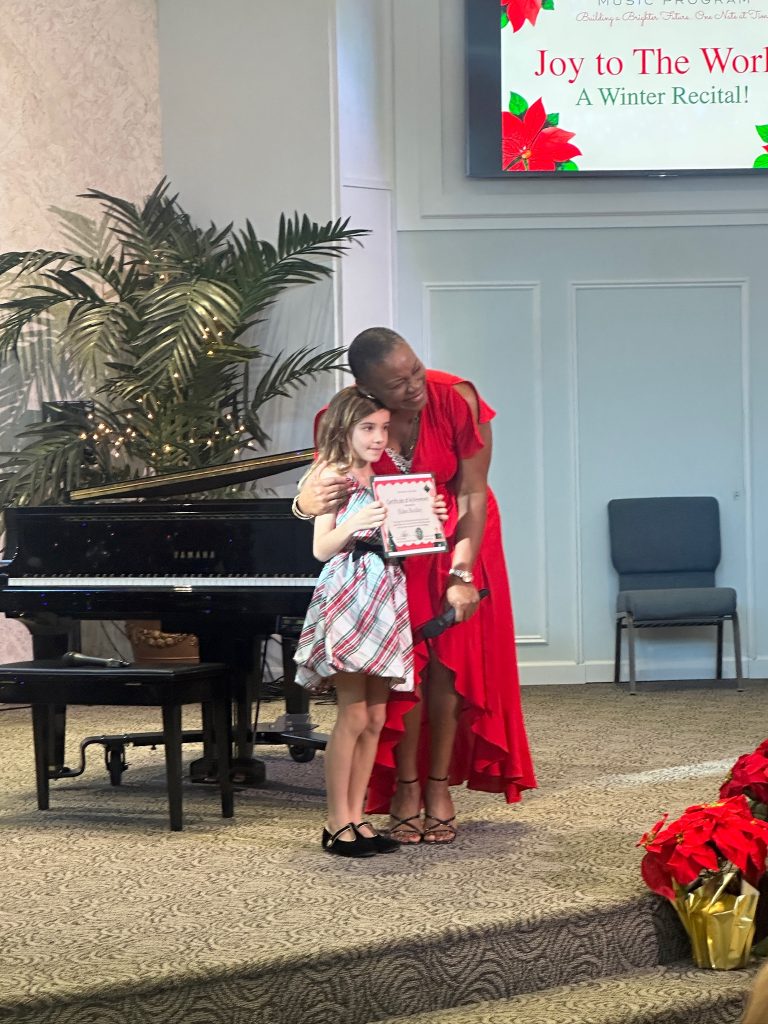 Vivia Taylor hugs a student holding a certificate in front of a piano at the 2025 Winter Recital.